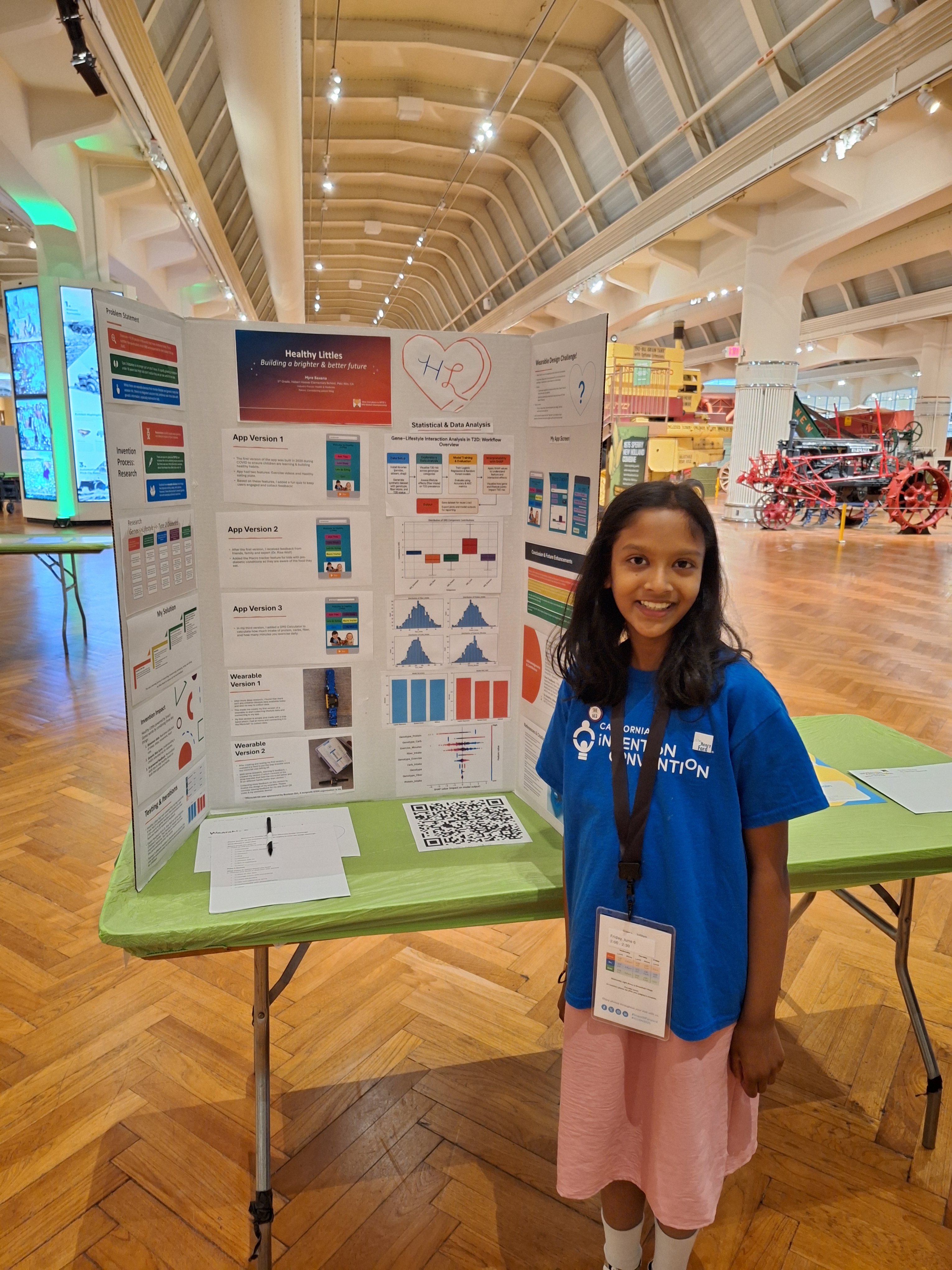 20250604_102357 A student in a blue t-shirt standing beside a tri-fold poster promoting her invention
